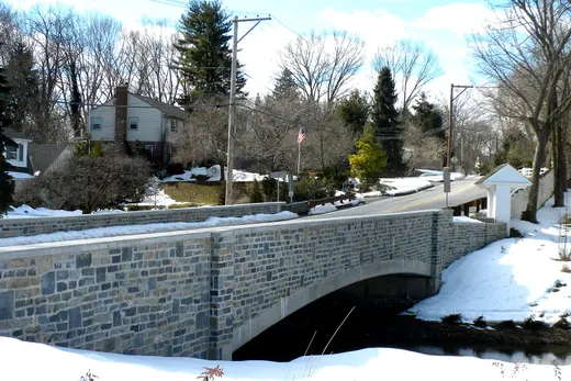 Stone bridge over stream with snowy roadside and houses, Tredyffrin Township, Pennsylvania
